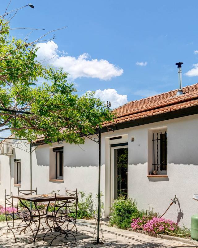 Cour arborée d'une maison basse aux murs blancs, table de jardin et barbecue