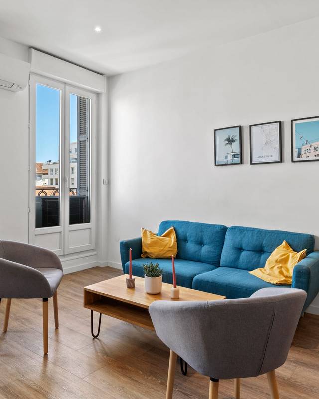 Salon clair avec canapé bleu, fauteuils gris et table basse en bois devant une porte-fenêtre